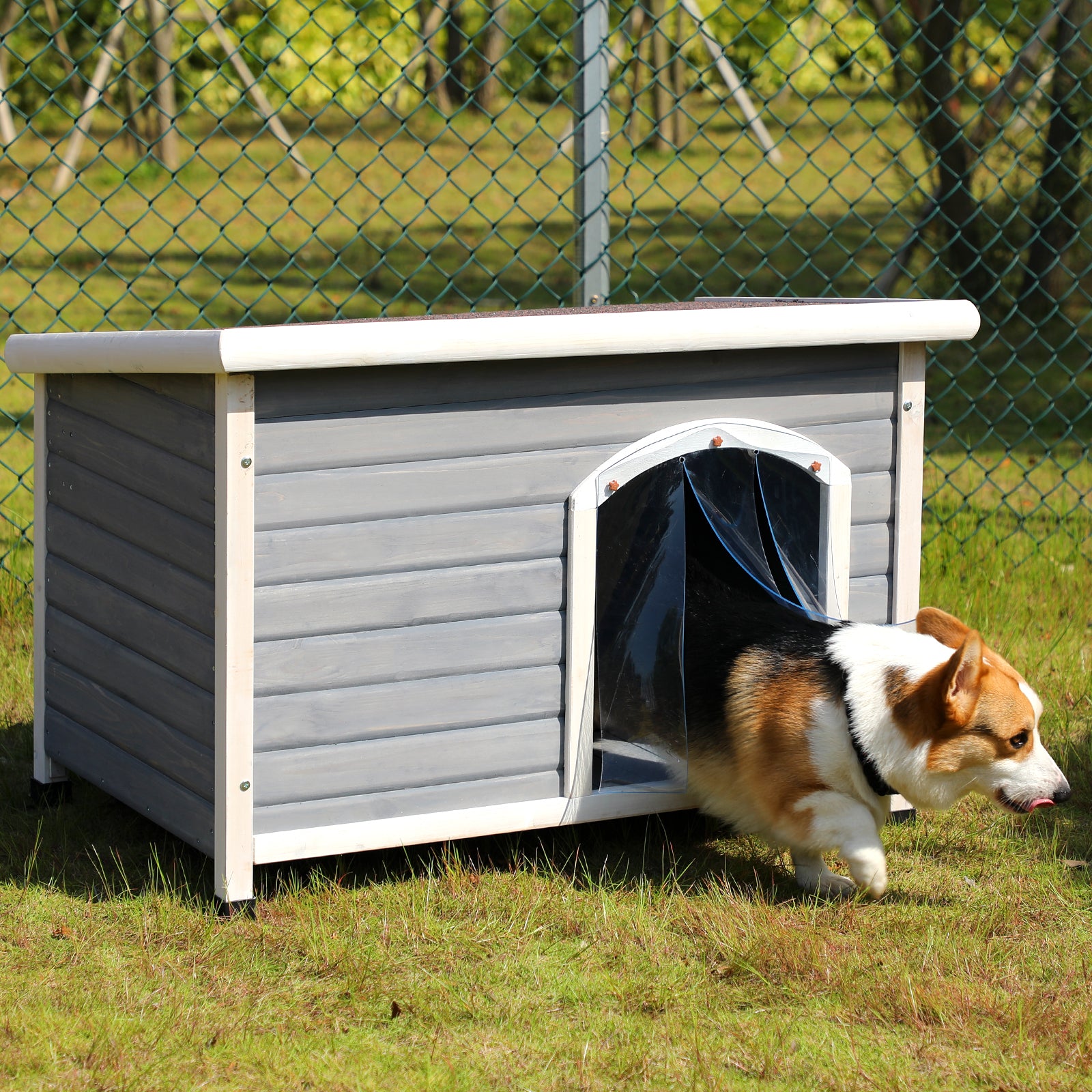 Dog entering a gray and white doghouse on grass with a chain-link fence in the background